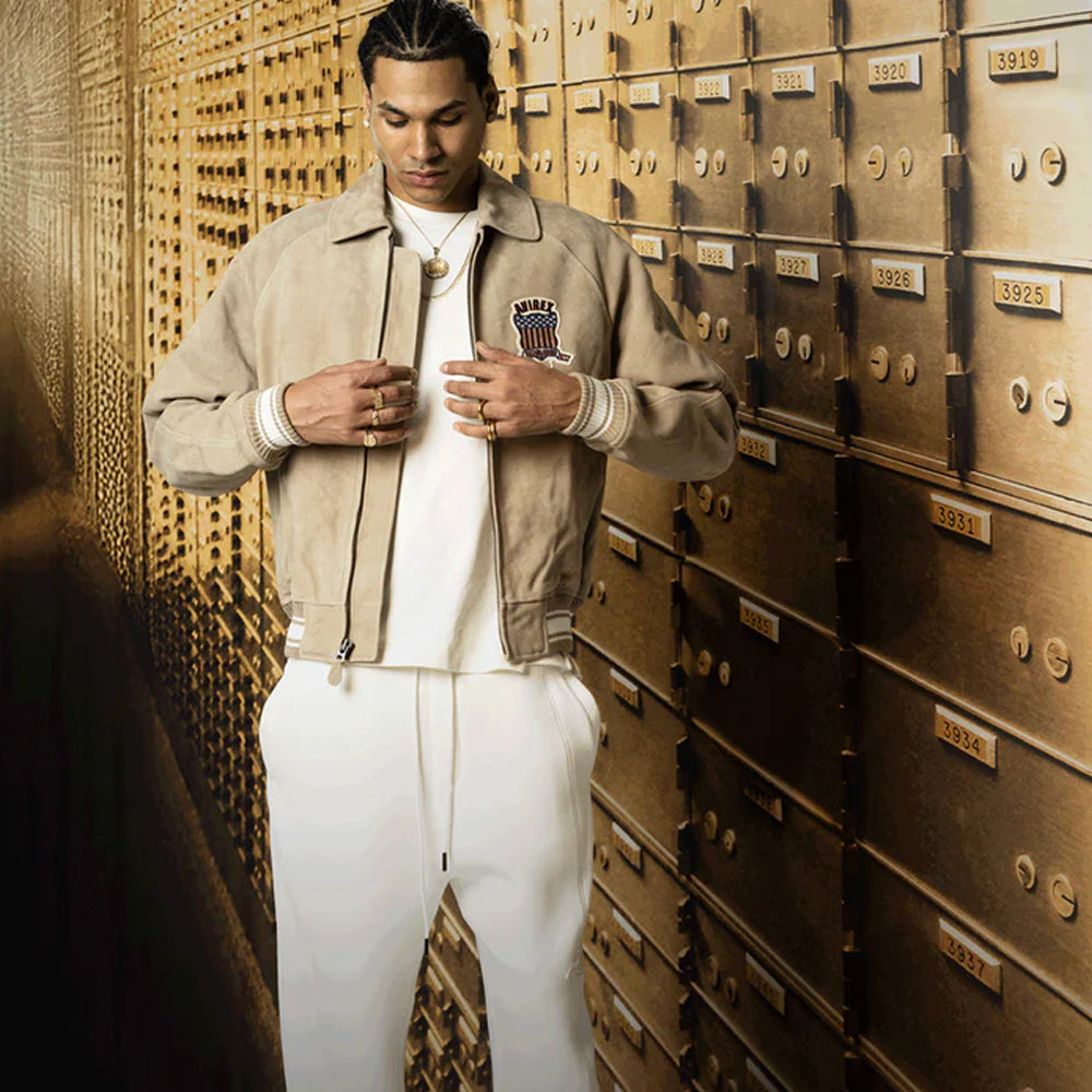 Man in beige jacket and white pants standing in front of a wall of gold safe deposit boxes.