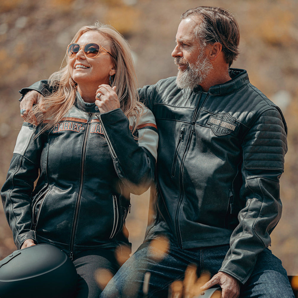 Two people wearing Harley Davidson jackets sitting together with a blurred natural background