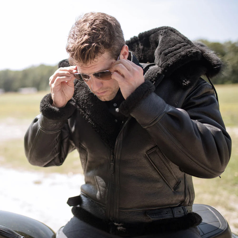 Man wearing men's midnight black sheepskin hooded aviator jacket with black fur ruff outdoors.
