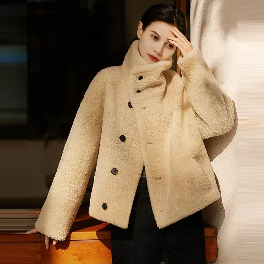 A woman standing up while fully buttoning the oversized plush shearling collar of her reversible leather coat for maximum wind protection.