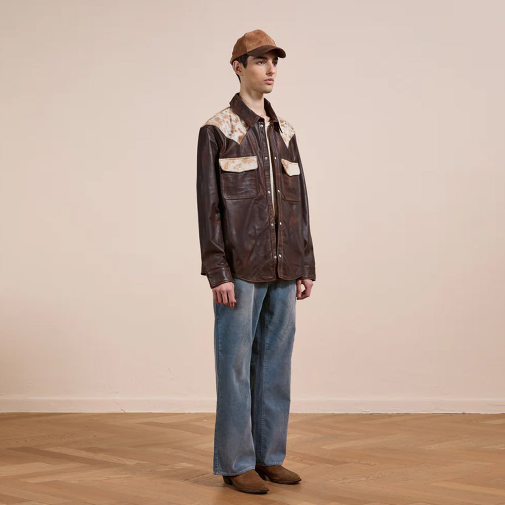 Person wearing a brown leather jacket with floral patterns and blue jeans on a plain background