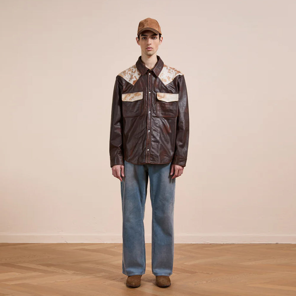 Man wearing a brown leather jacket with patterned accents, blue jeans, and a cap against a plain background.