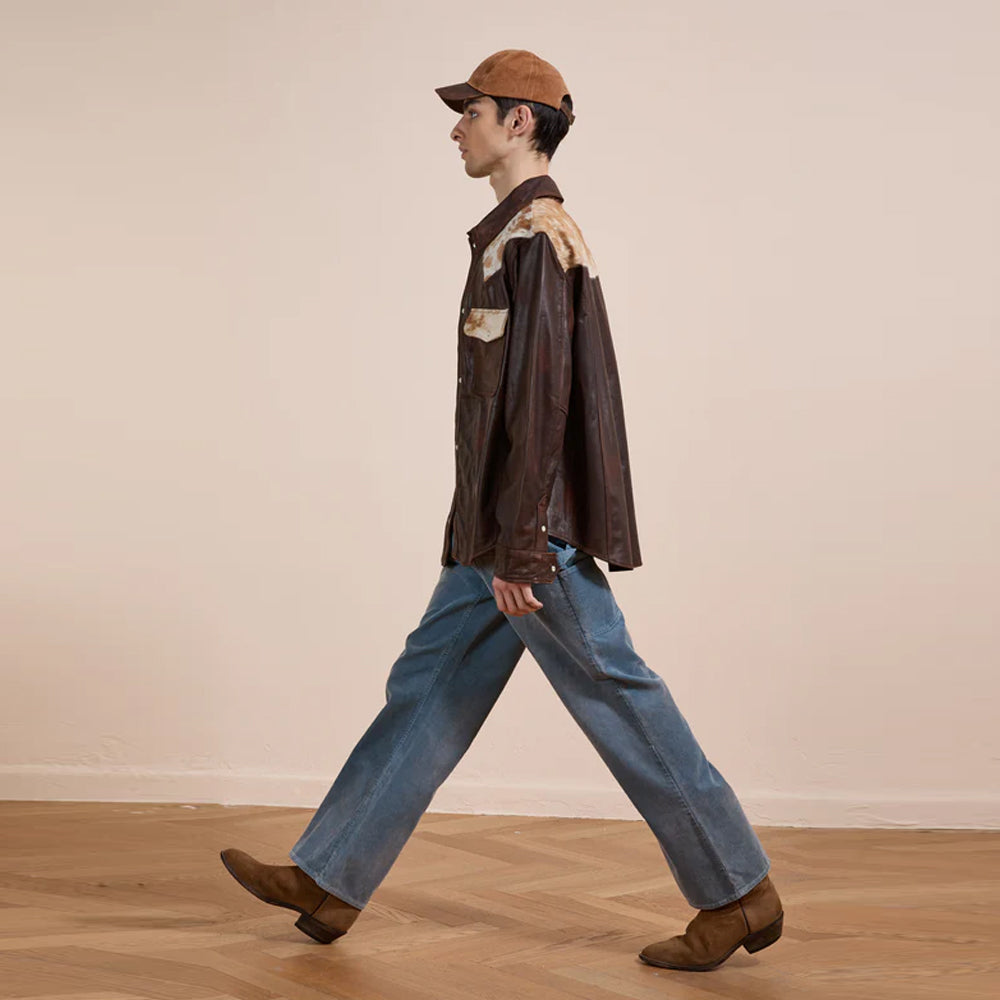 Person wearing a brown jacket, blue jeans, and a cap walking on a wooden floor.