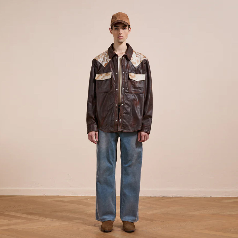 Person wearing a brown leather jacket with floral patterns and blue jeans on a plain background