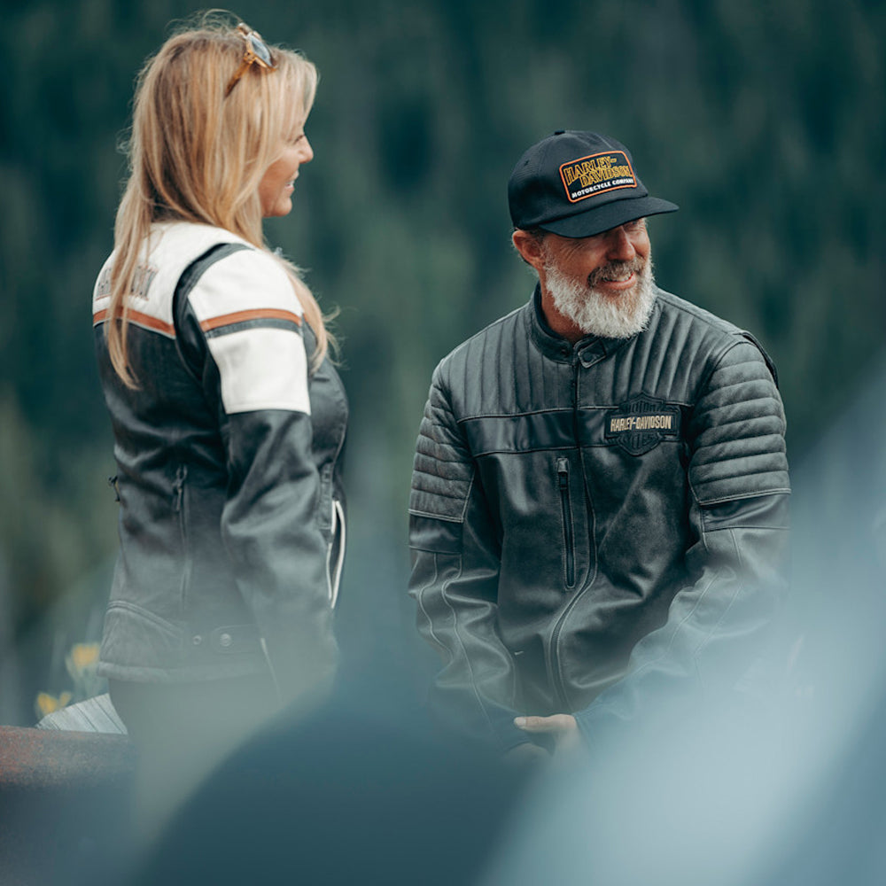 Two people wearing Harley-Davidson jackets and hats in a forest setting