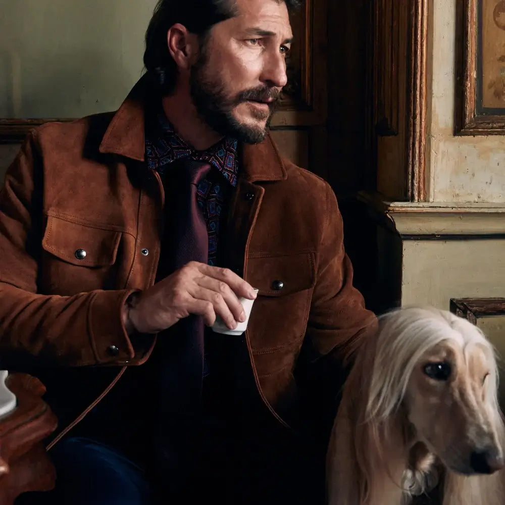 Man in a brown suede leather jacket sitting outdoors with a dog, holding a tea cup, and enjoying a relaxed moment.