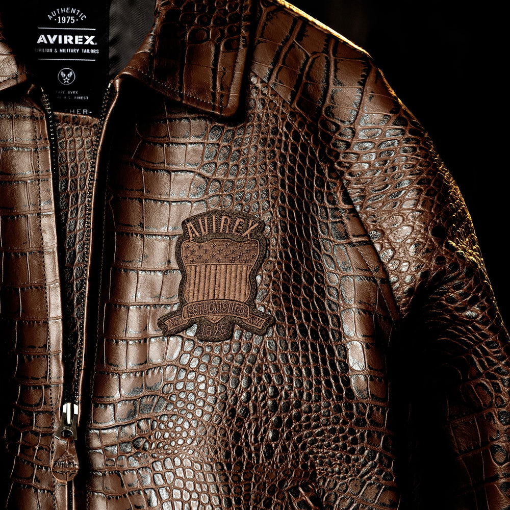 Campaign close-up highlighting espresso croc embossing, point collar, front zipper and tonal Avirex crest