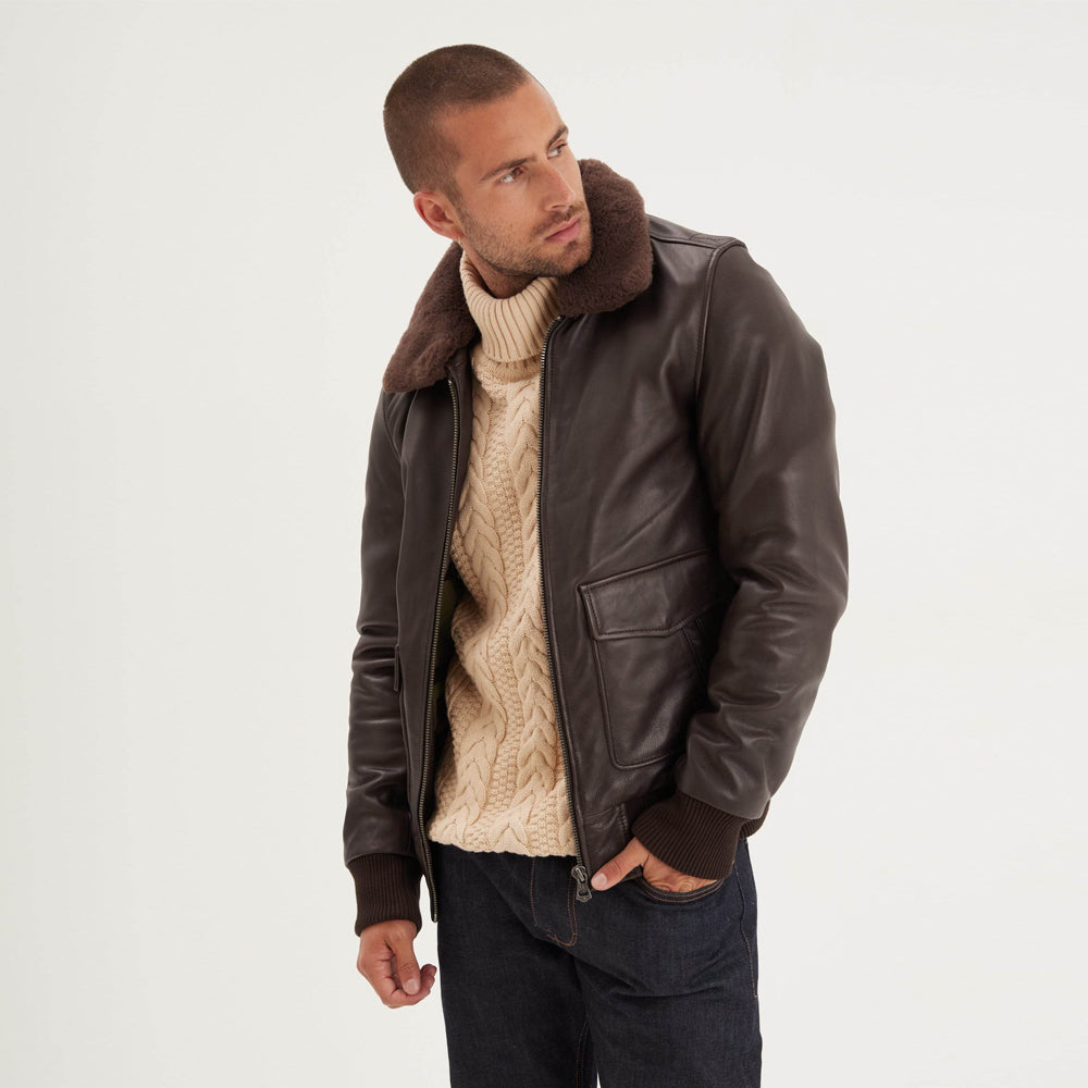 Three-quarter front view of brown leather flight bomber layered over a cream cable-knit turtleneck and dark denim.