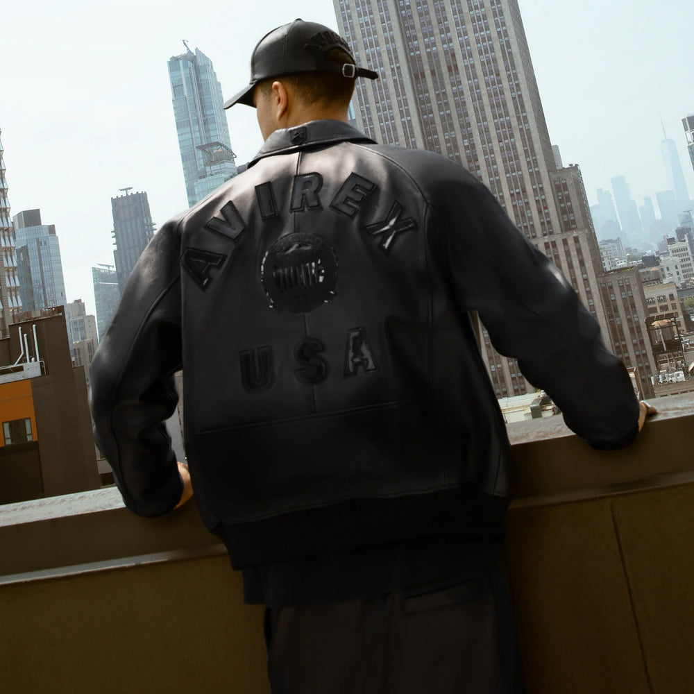 Rooftop rear campaign view of men’s black Avirex USA leather bomber displaying tonal raised back lettering