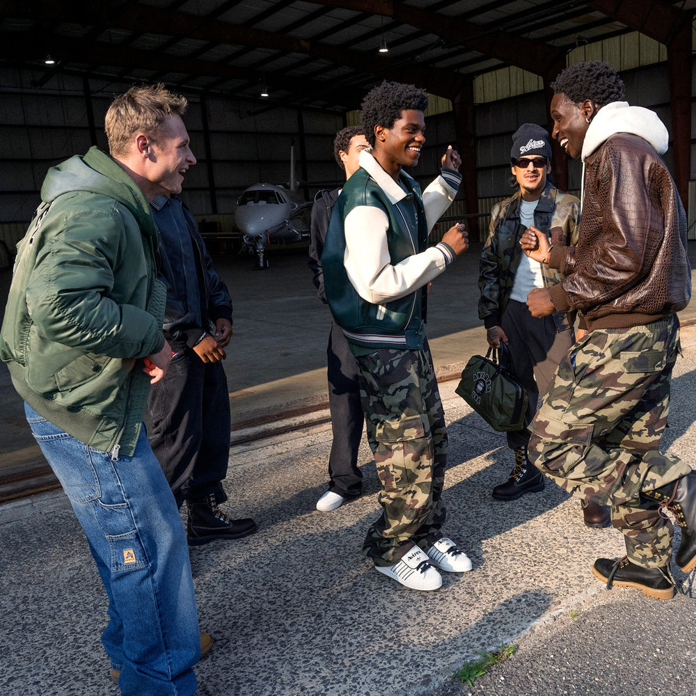 Men’s Avirex brown flight-inspired jacket styled with camouflage trousers in a group hangar scene