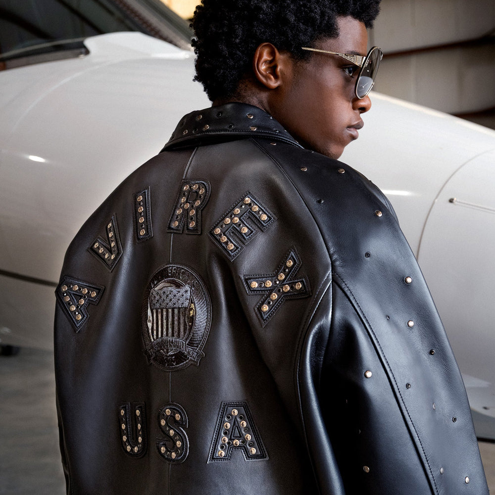 Campaign rear view of a man wearing the black Avirex Studded Icon jacket beside an aircraft