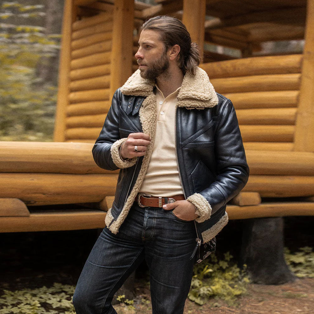 Male model standing near wooden log cabin wearing open men's black aviator shearling coat with beige fleece lining