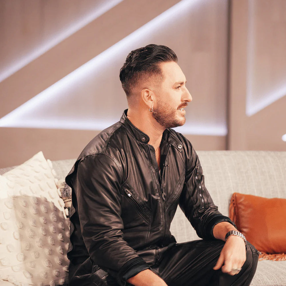 Seated model wearing a men's full-body black leather catsuit with long sleeves and utility pockets