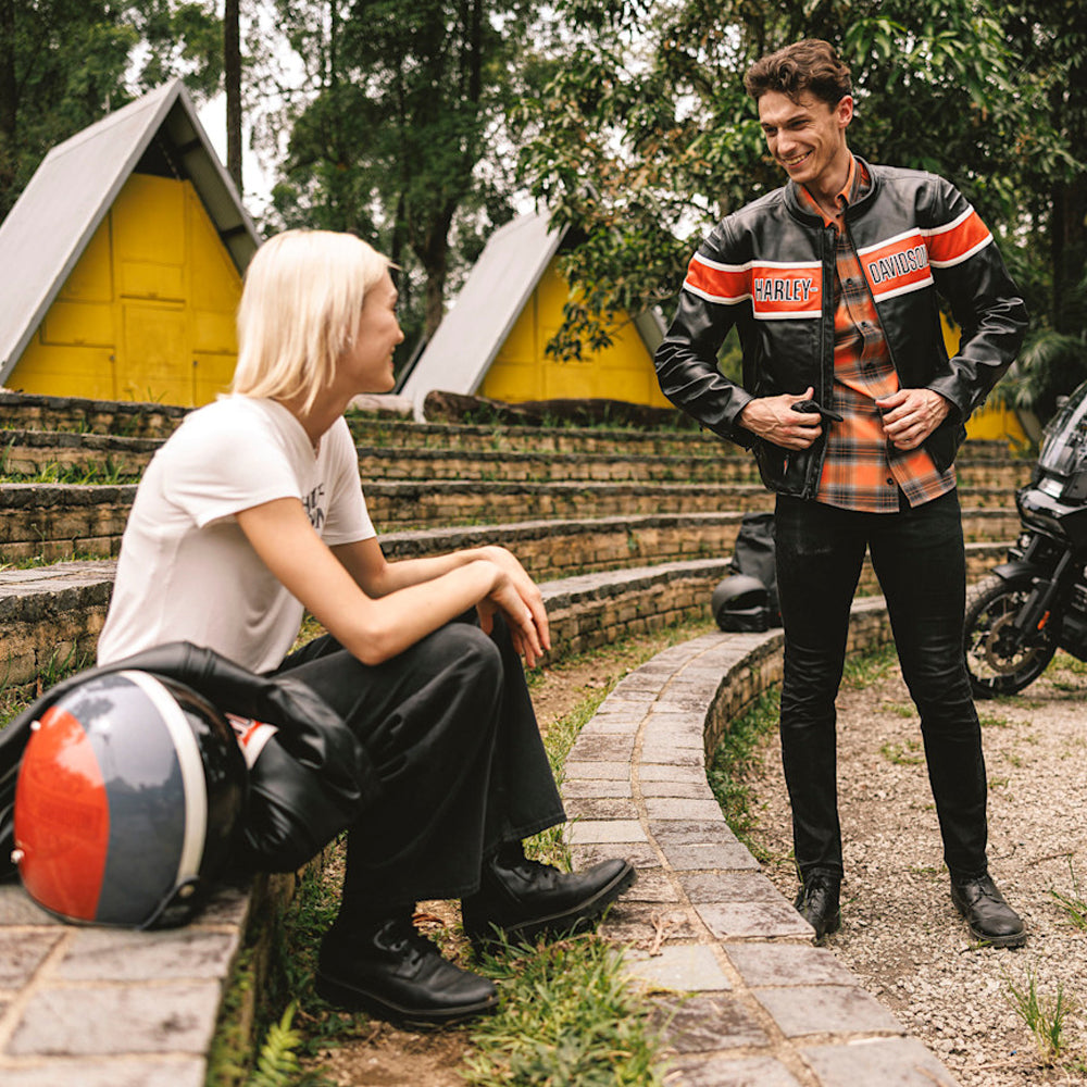 Man wearing a Harley-Davidson-style black leather biker jacket beside a woman and motorcycles at an outdoor campsite