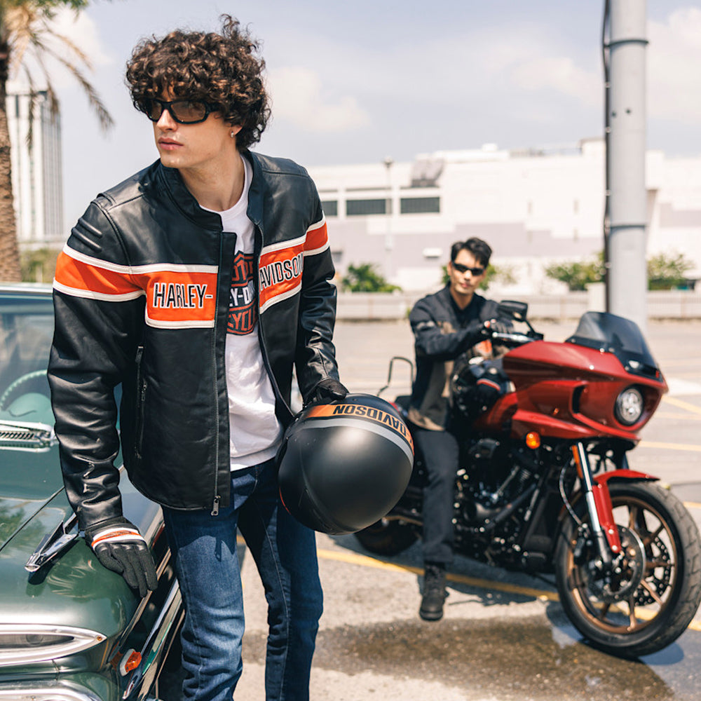 Man in a black Harley-Davidson-style motorcycle leather jacket holding a helmet beside a vintage car and red touring bike