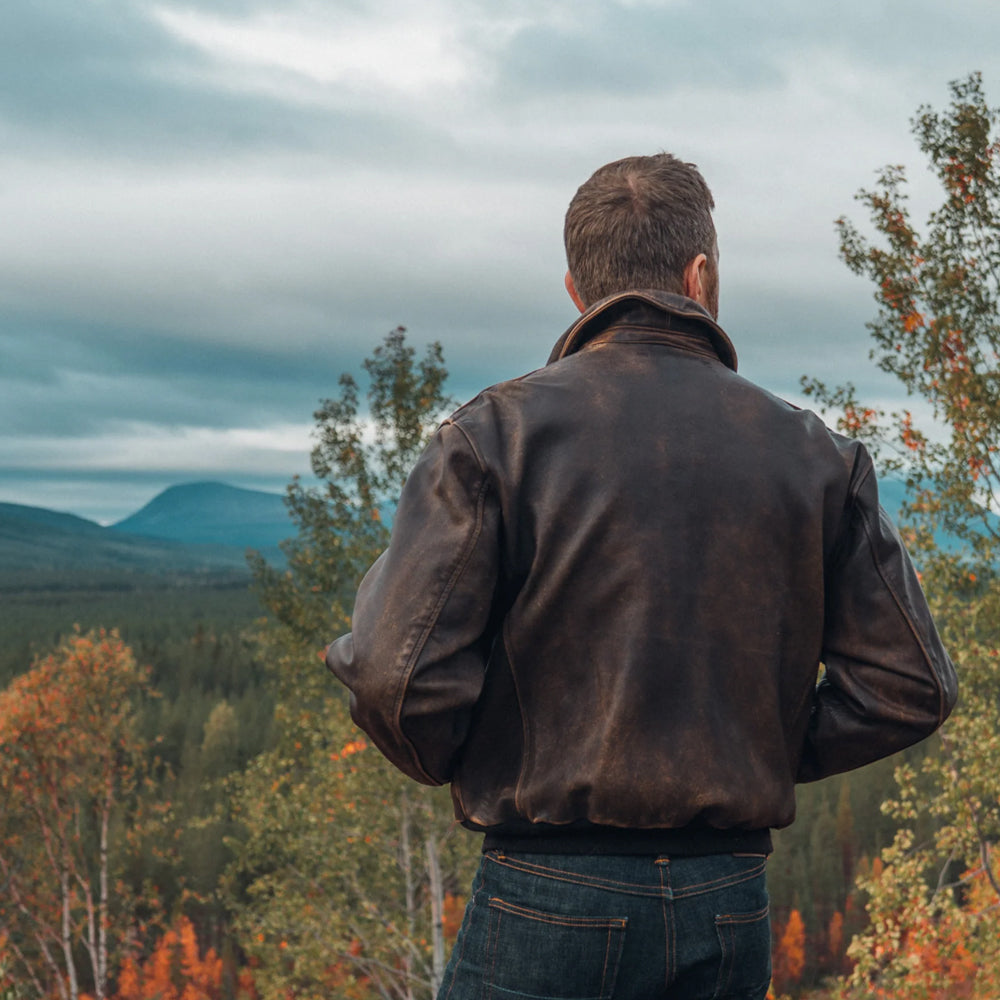 Rear outdoor shot of male model wearing heritage full-grain brown aviator jacket overlooking mountain forest.