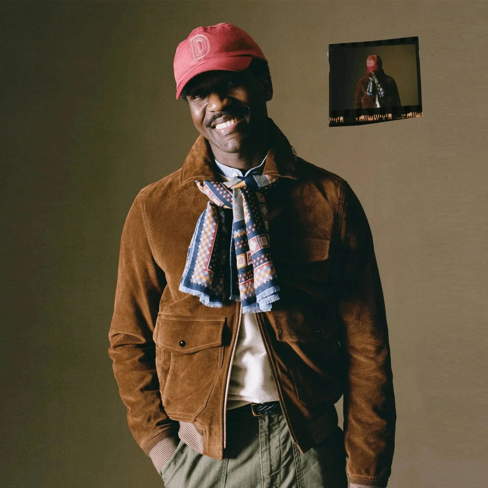Men's tobacco suede bomber styled with an olive trouser, patterned scarf and cap in a warm indoor setting.