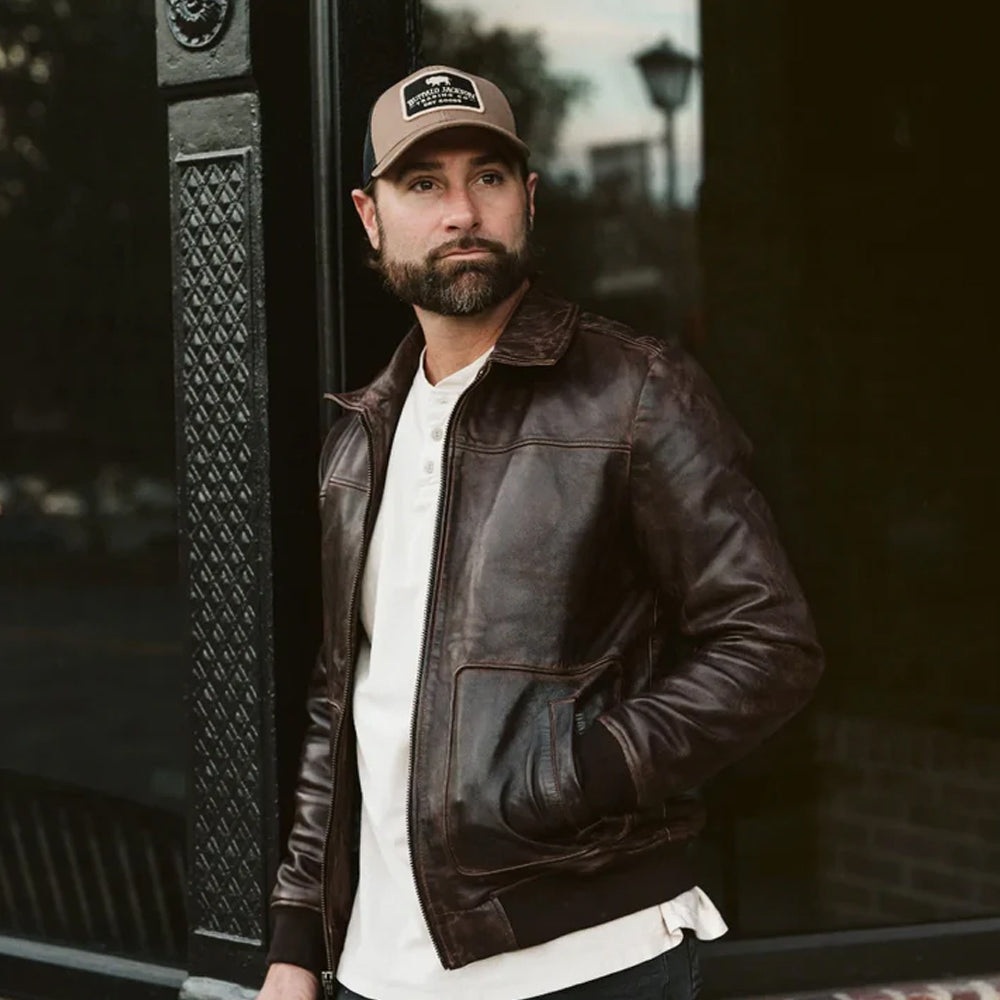 A man wearing a dark brown vintage A2 leather bomber jacket with a brown cap, standing outdoors against a dark window frame.