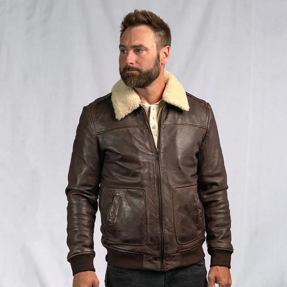 A bearded man modeling a rugged brown distressed leather aviator bomber jacket with a soft cream shearling collar against a clean studio backdrop.