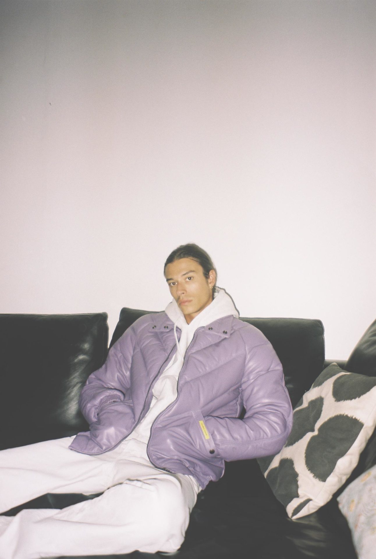 Lifestyle view of a man wearing the lilac down jacket sitting on a sofa with hands in his side pockets.