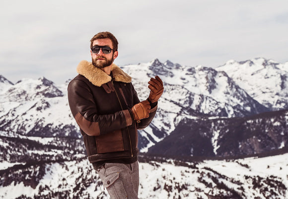 Man wearing brown shearling leather jacket with fur collar in snowy mountains - genuine sheepskin cold weather protection