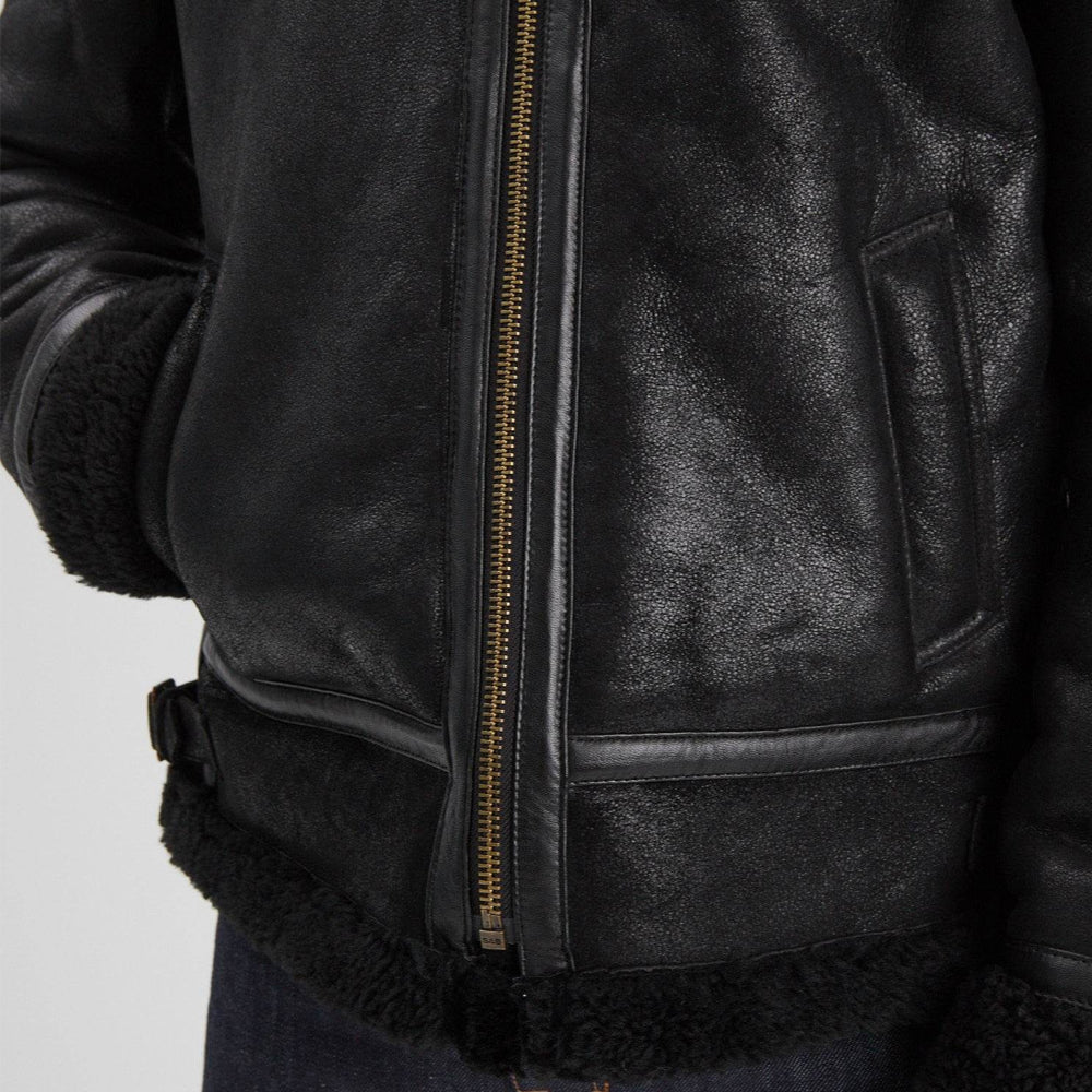 Black sheepskin aviator jacket detail showing open slanted welt pocket, leather-bound zipper, and shearling hem