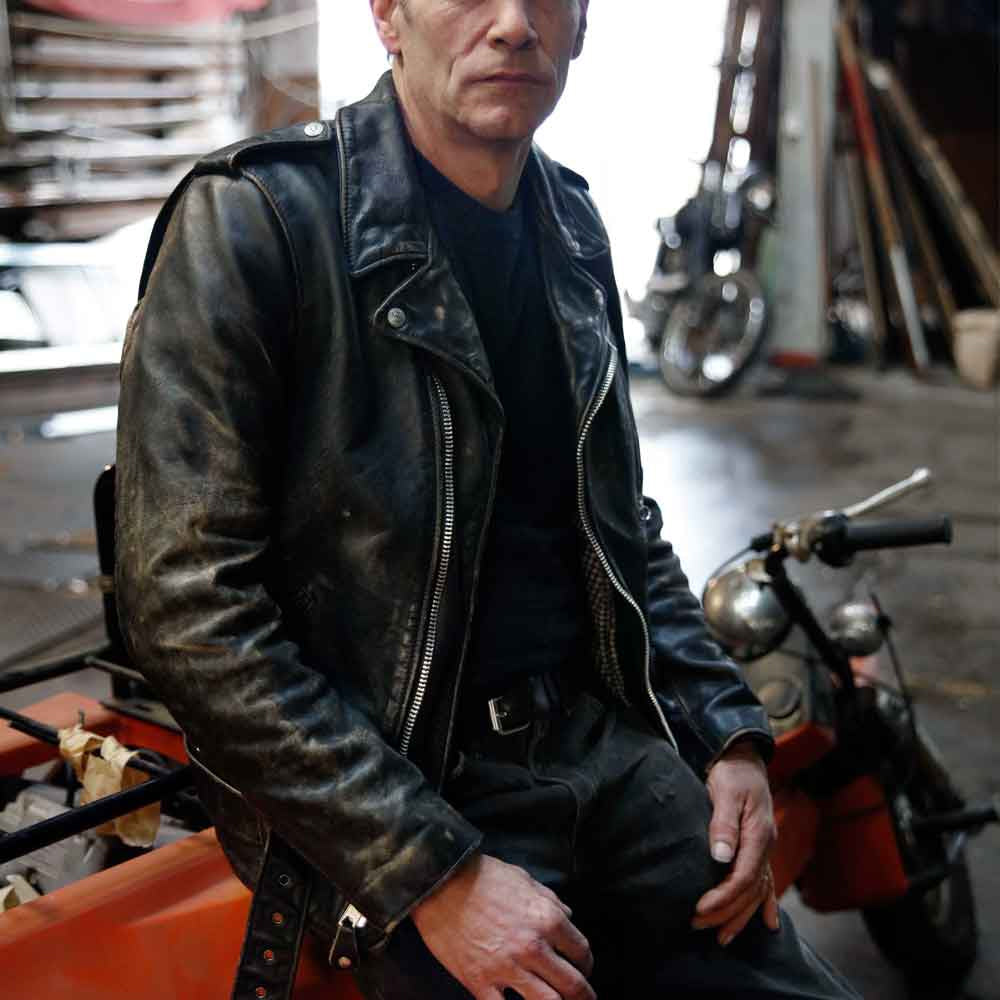 Model wearing waist-length vintage distressed leather motorcycle jacket with epaulettes, asymmetrical front and adjustable belt