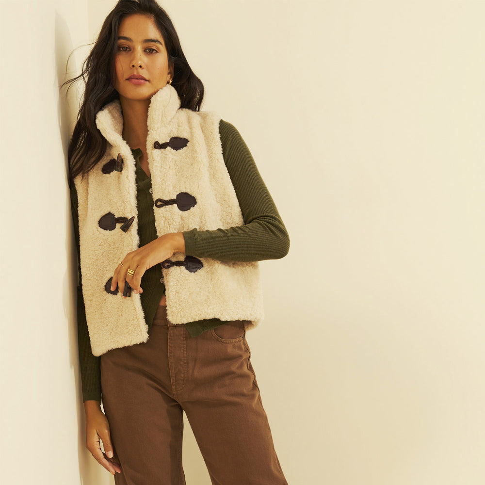 Woman wearing a cream sherpa toggle vest over an olive ribbed top, front view.