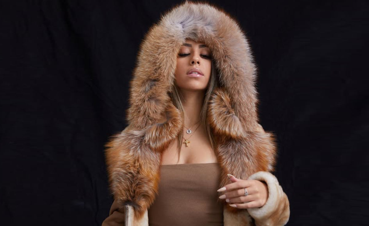 Woman wearing a brown luxury shearling jacket with a thick real fox fur trim hood against a dark background.